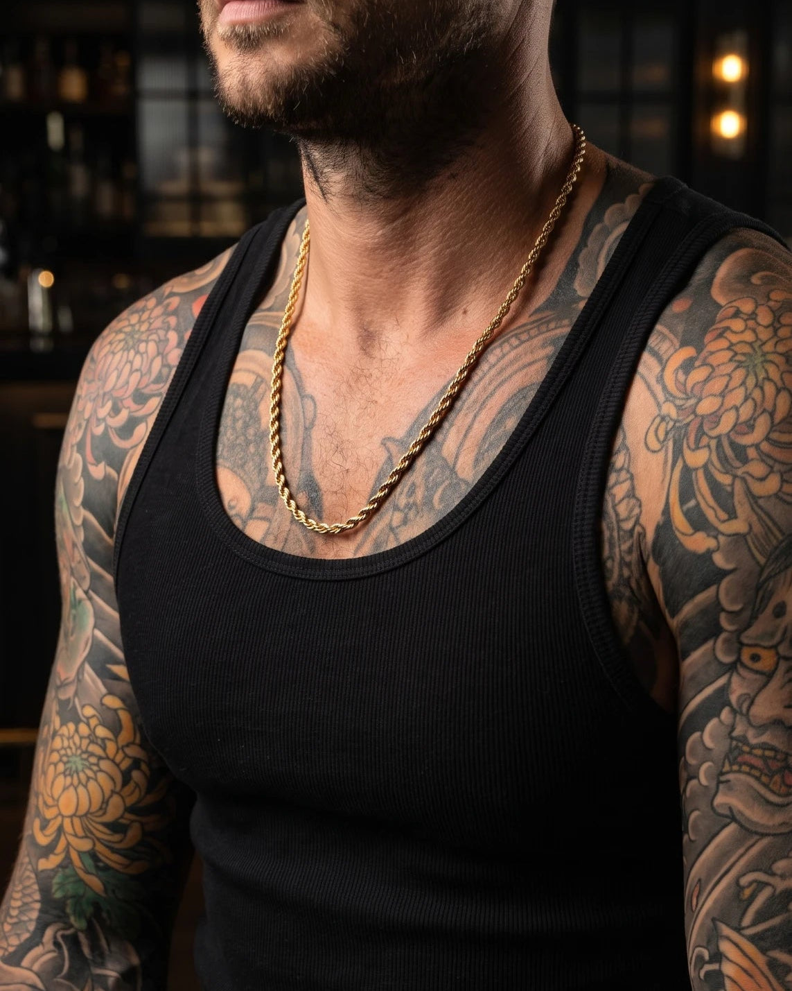 A close-up of a man wearing a black tank top and a chunky gold chain necklace, with detailed tattoos visible on his arms and chest, set against a dark, softly blurred background.