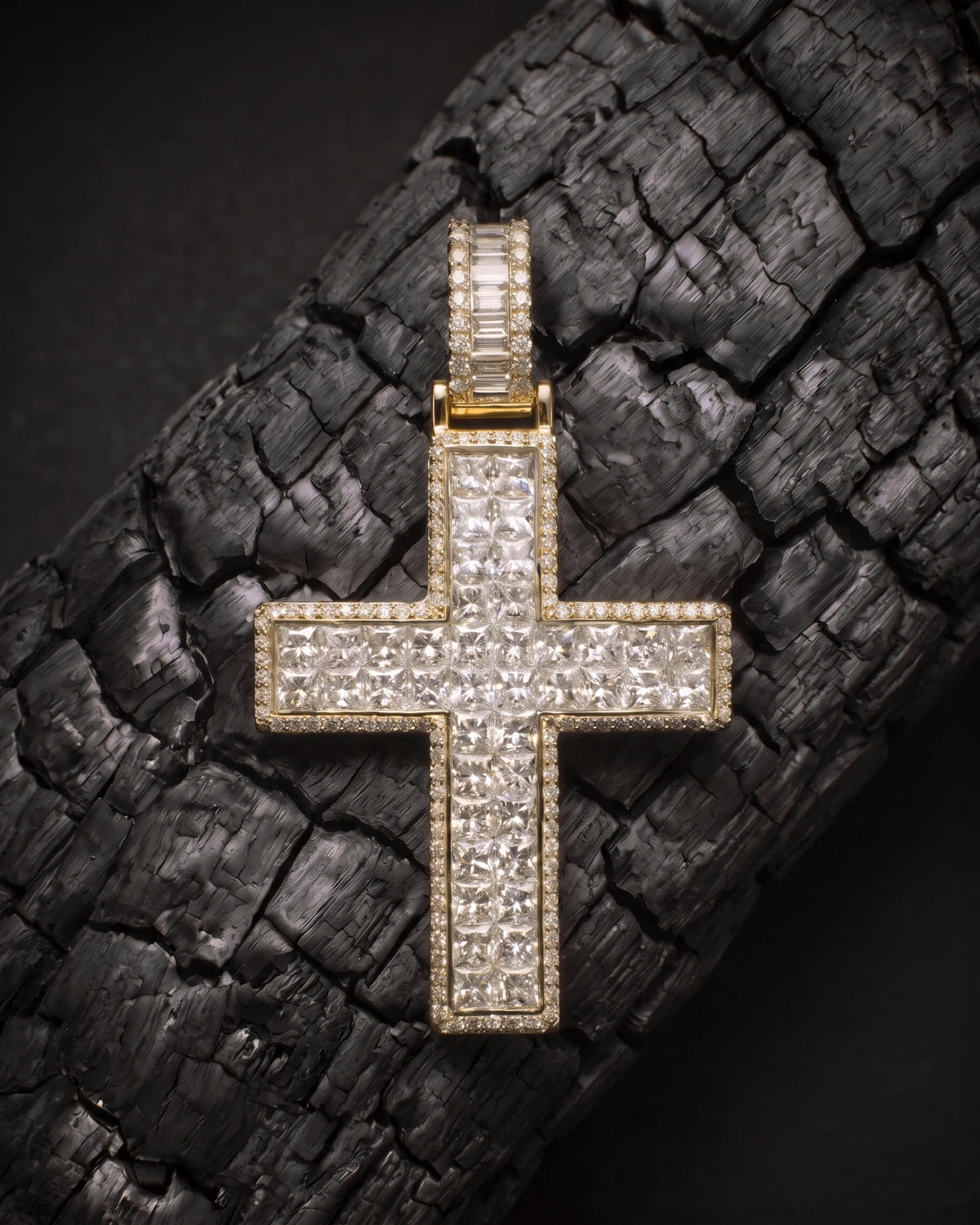 The Sanctum Cross in 14K yellow gold resting on charred wood, invisible-set princess diamonds with pave border and baguette bail